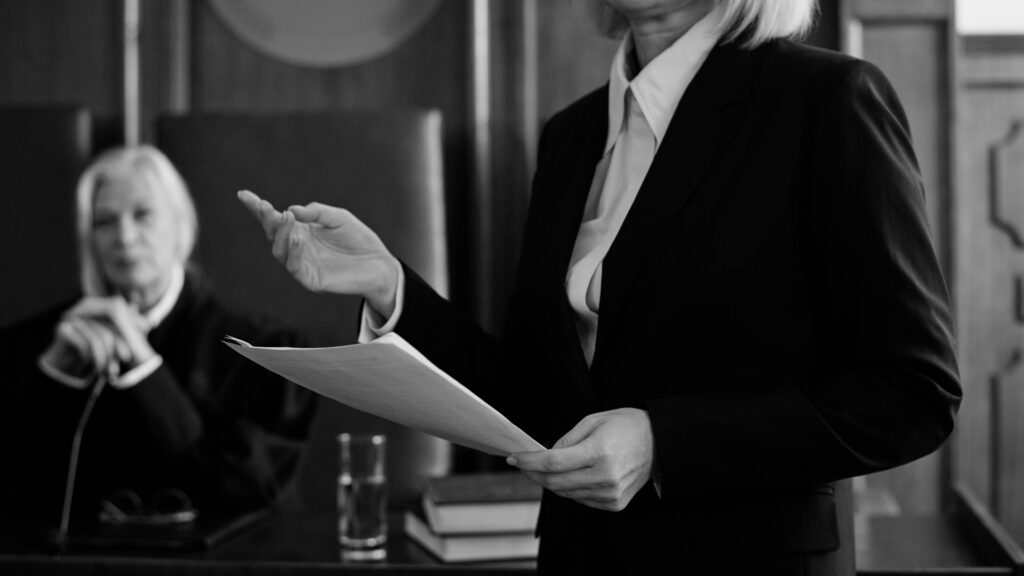 Attorney standing and speaking while holding documents in a conference room, with another legal professional seated nearby.