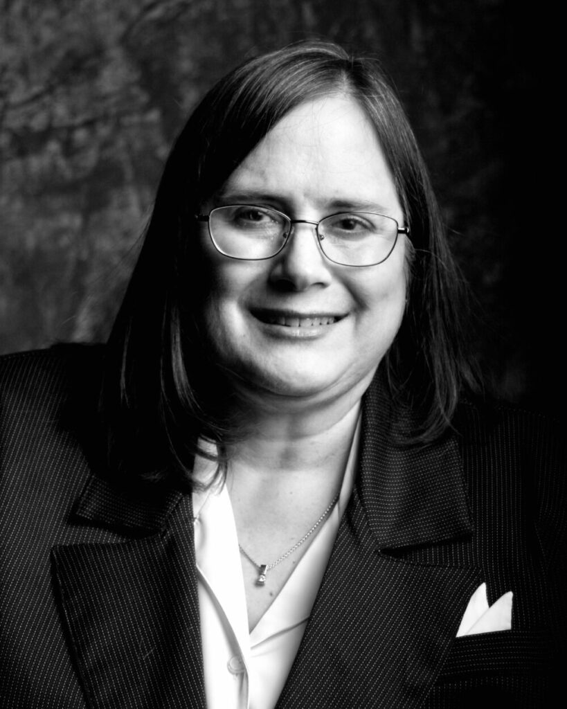 Professional black-and-white headshot of Heidi Hagen wearing glasses and a blazer, smiling in front of a dark textured background.